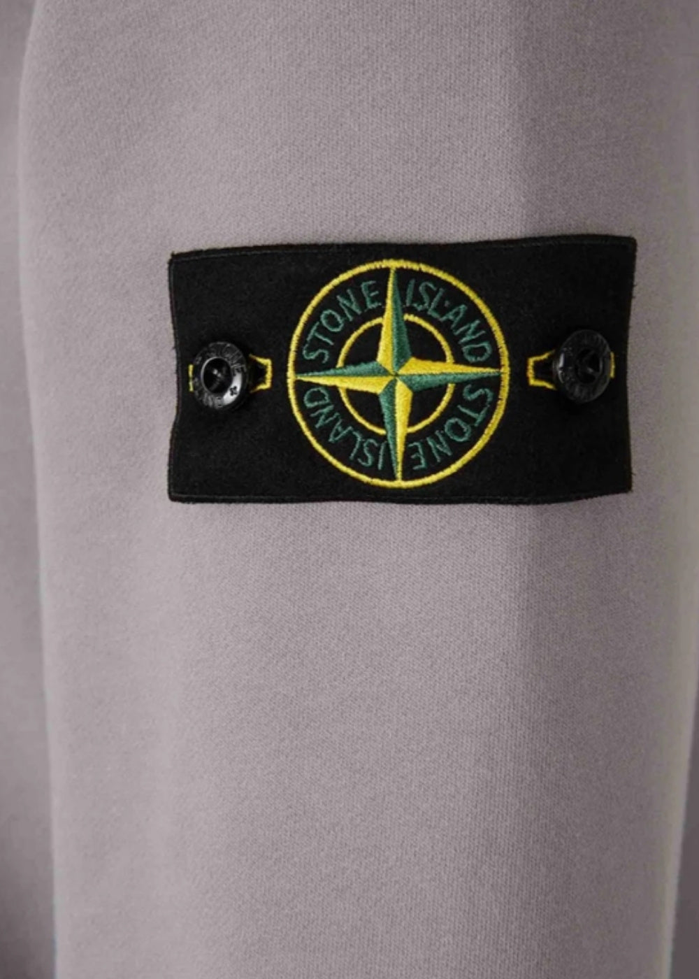 Gros plan sur le badge boussole Stone Island noir fixé par deux boutons sur la manche gauche d'un sweat-shirt gris.