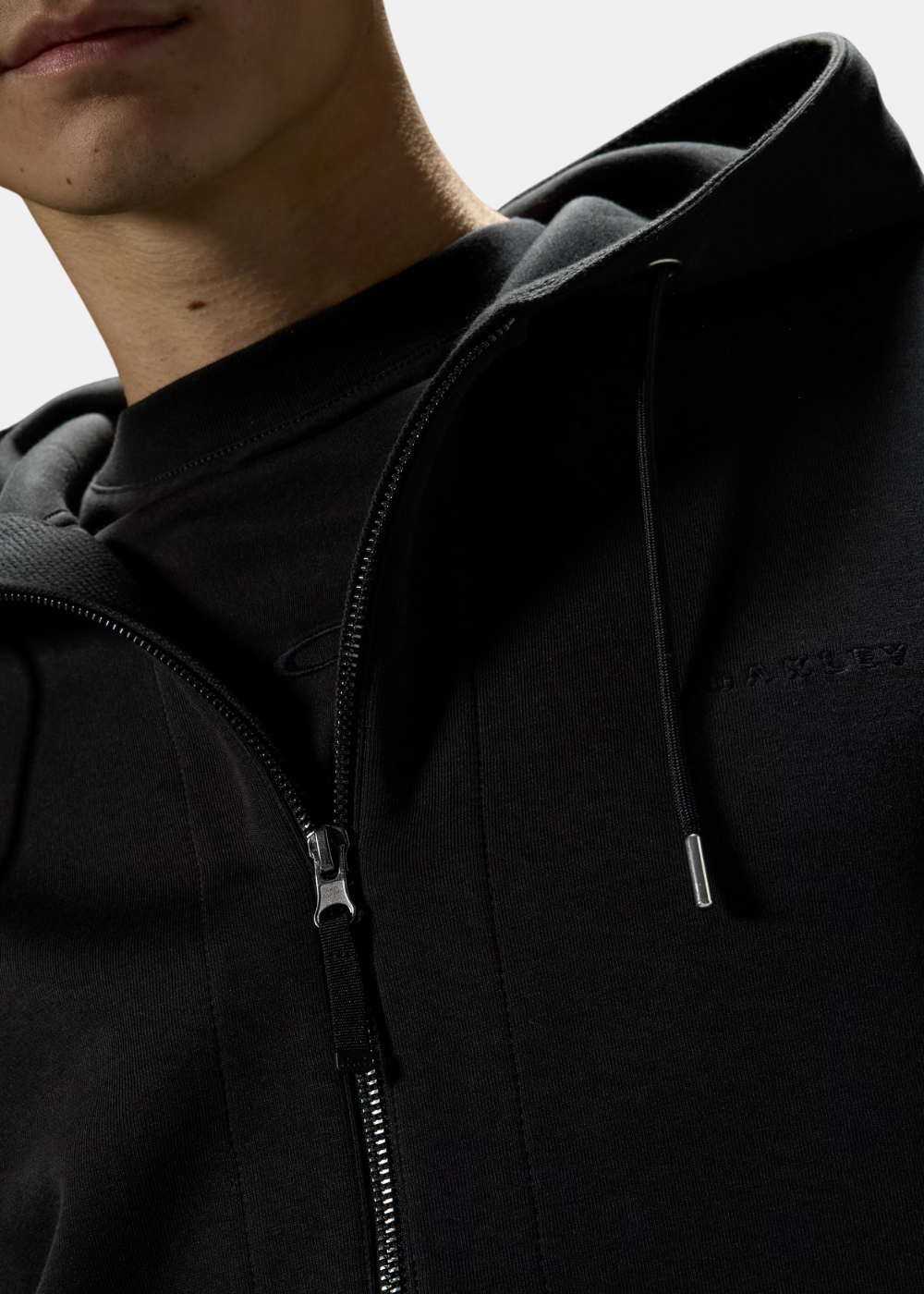 Zoom porté sur la fermeture éclair métallique et le logo Oakley discret brodé sur la poitrine du sweat noir.