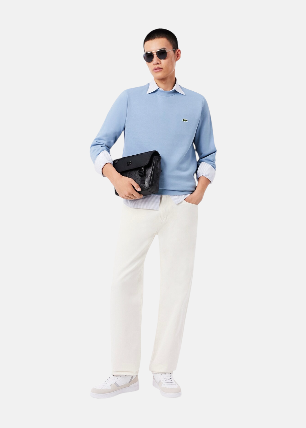 Tenue complète avec sweatshirt Lacoste bleu, pantalon blanc, baskets et sacoche noire portée par un mannequin.