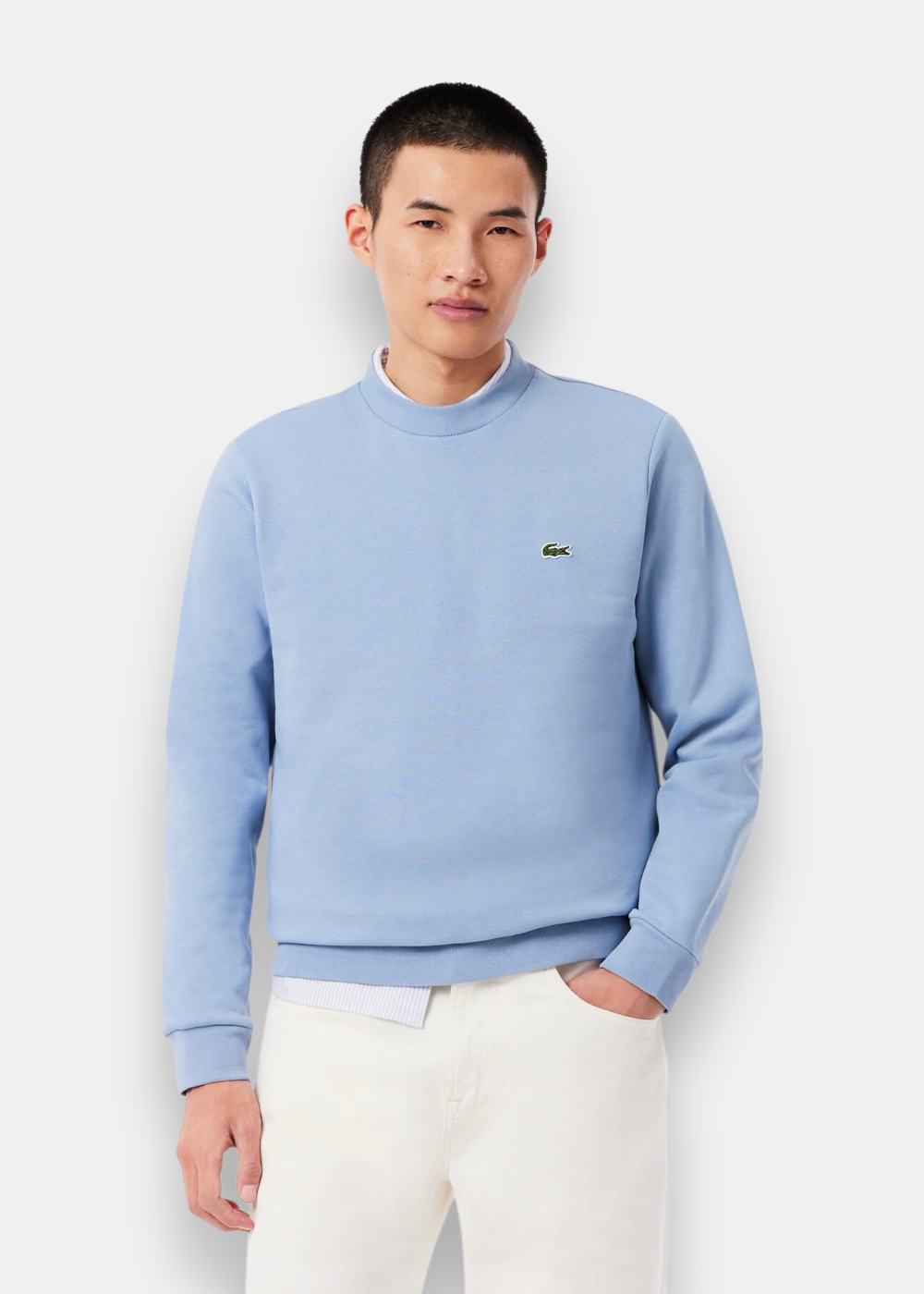 Homme portant le sweatshirt Lacoste bleu clair avec un col rond sur une chemise rayée et un pantalon blanc.