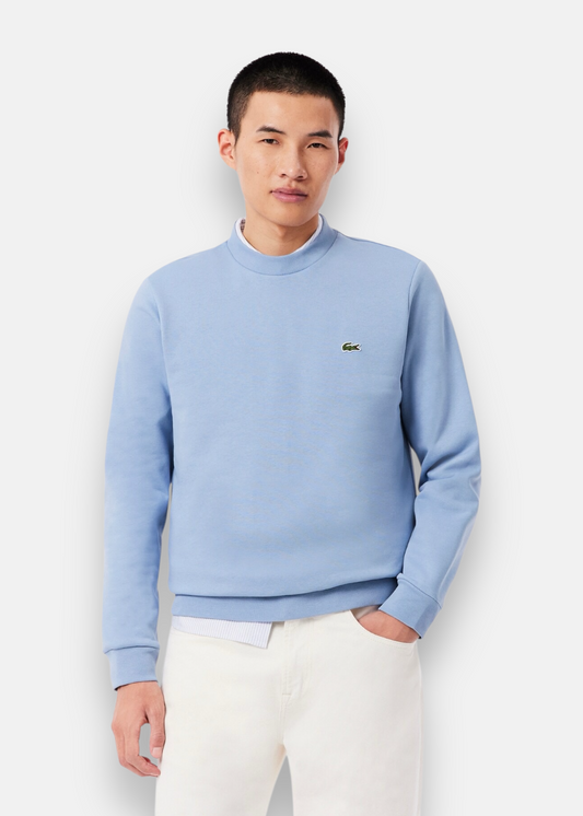 Homme portant le sweatshirt Lacoste bleu clair avec un col rond sur une chemise rayée et un pantalon blanc.