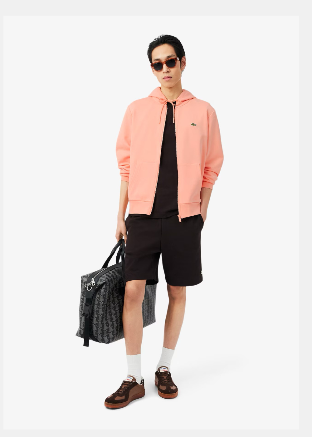 Silhouette complète avec sweatshirt rose corail Lacoste, short noir, lunettes de soleil et sac de sport