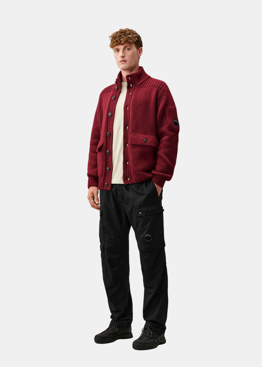 Mannequin homme portant le t-shirt beige C.P. Company sous un cardigan en grosse maille rouge bordeaux à col montant, pantalon cargo noir et baskets
