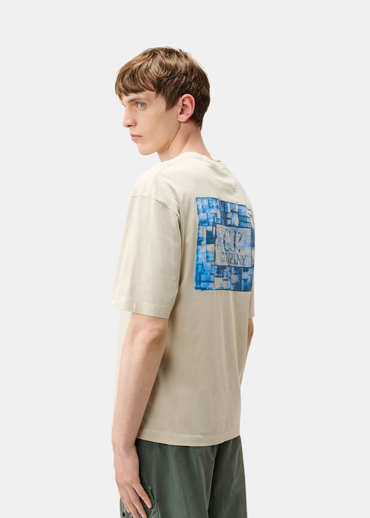 Vue de trois-quarts dos d'un t-shirt beige C.P. Company porté mettant en valeur le grand imprimé logo bleu.