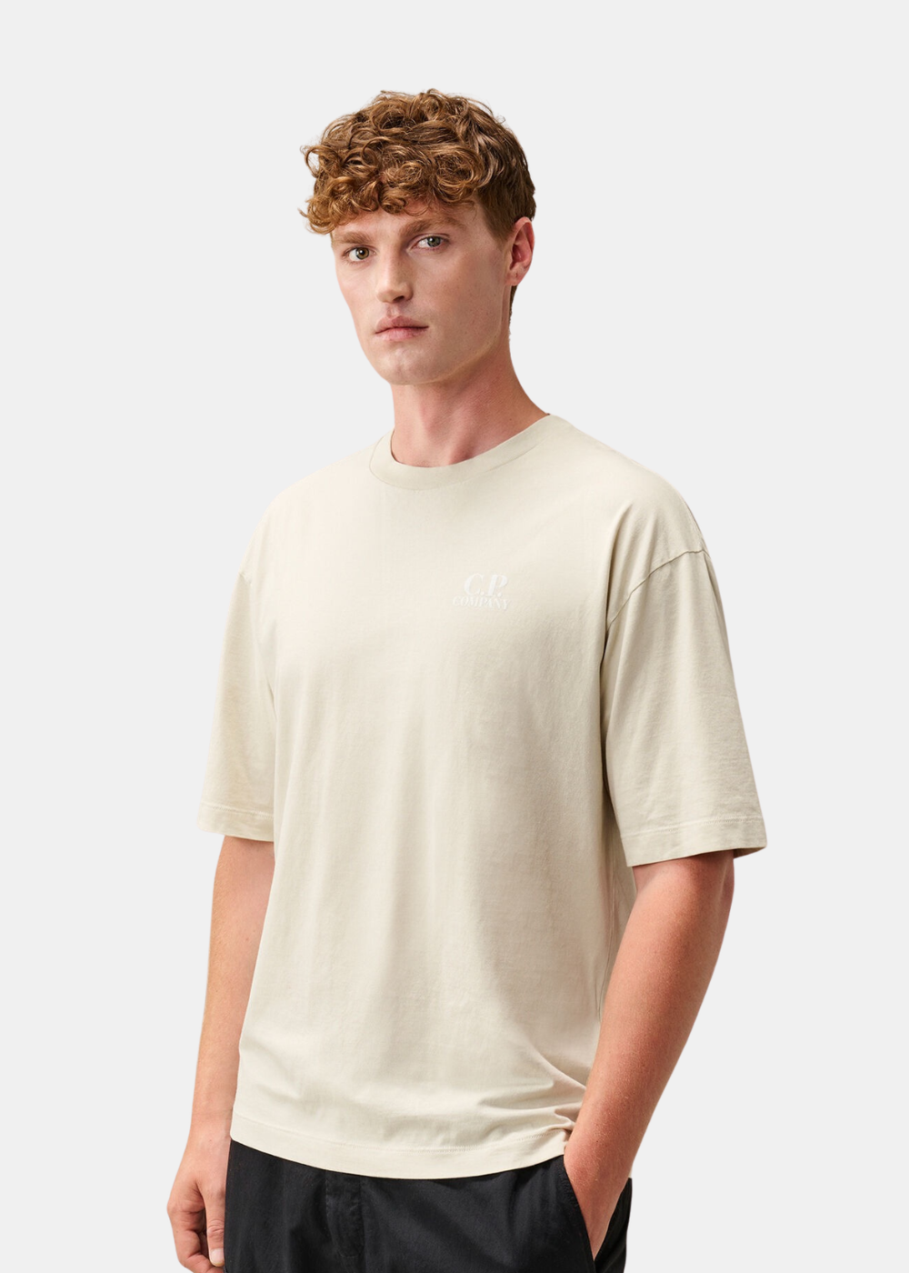 Mannequin homme de trois-quarts portant le t-shirt beige C.P. Company col rond avec logo blanc sur le torse et pantalon noir