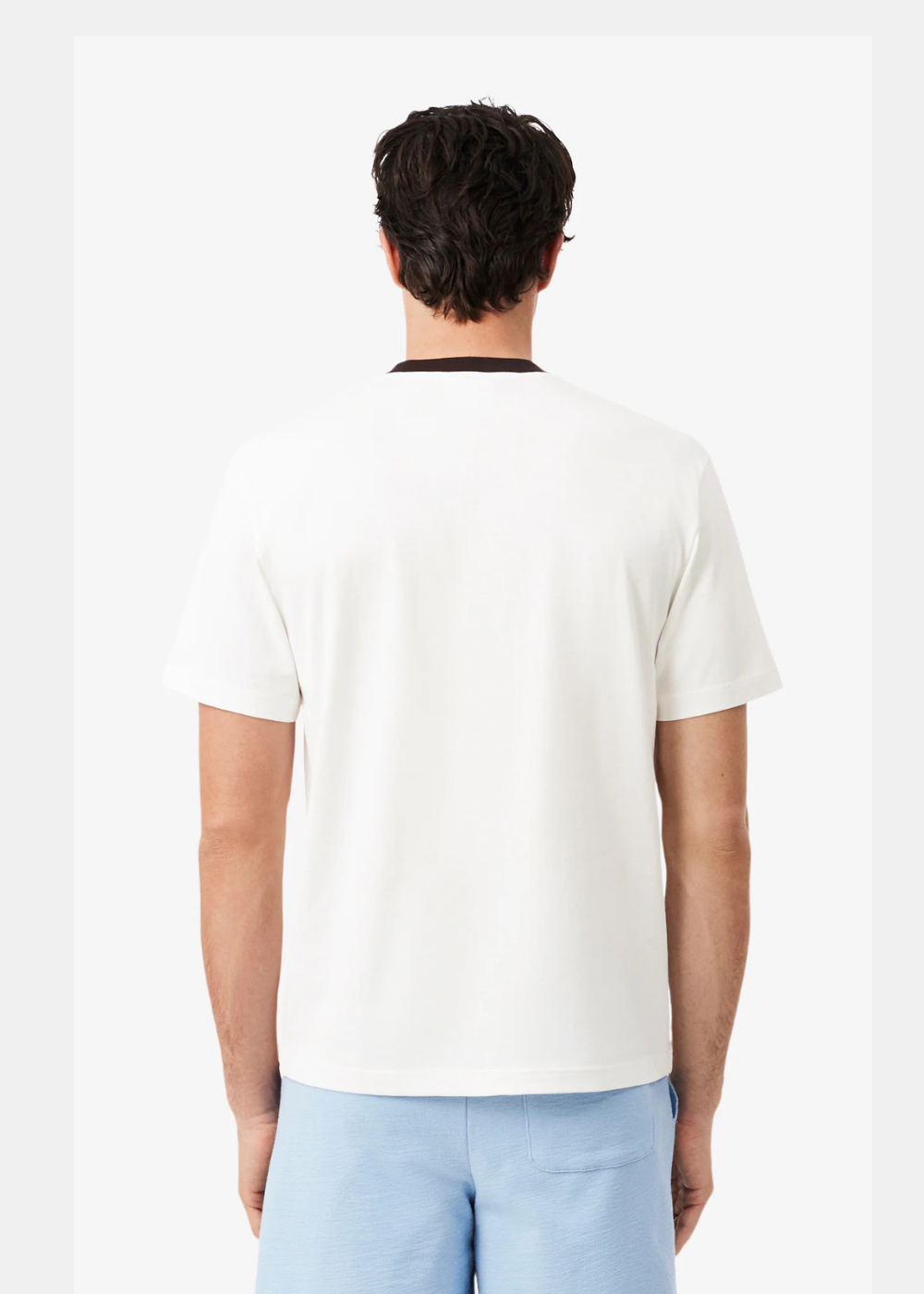 Vue de dos d'un homme portant le t-shirt Lacoste blanc à col noir et un short bleu ciel sur fond neutre.