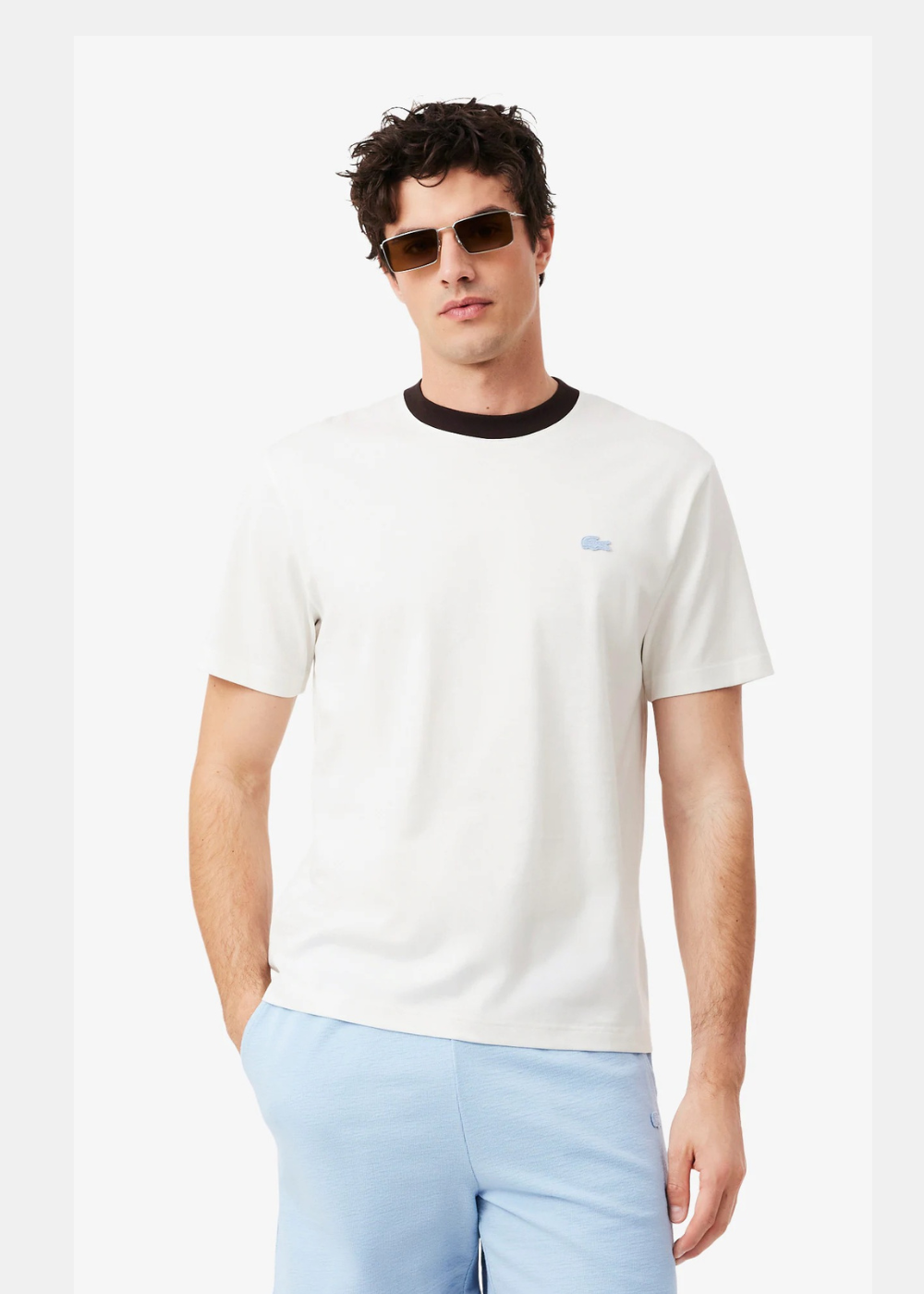 T-shirt Lacoste blanc porté par un homme avec un short bleu ciel et des lunettes de soleil. Vue de face sur fond blanc.