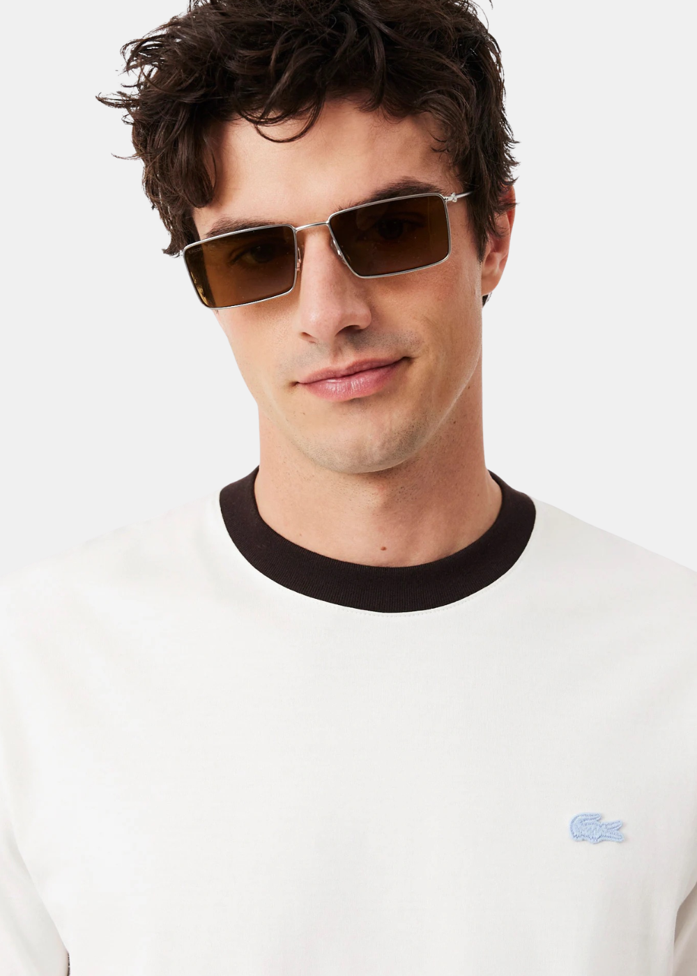 Gros plan sur le haut d'un t-shirt Lacoste blanc, mettant en avant le col noir contrasté et le logo crocodile bleu ciel brodé.