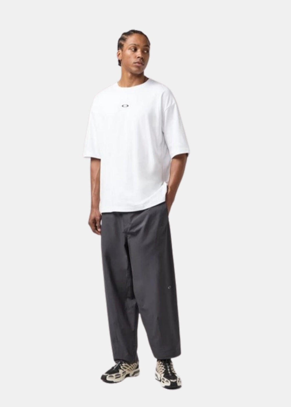 Porté en pied d'un t-shirt blanc Oakley ample associé à un pantalon gris large et des sneakers, silhouette streetwear décontractée.