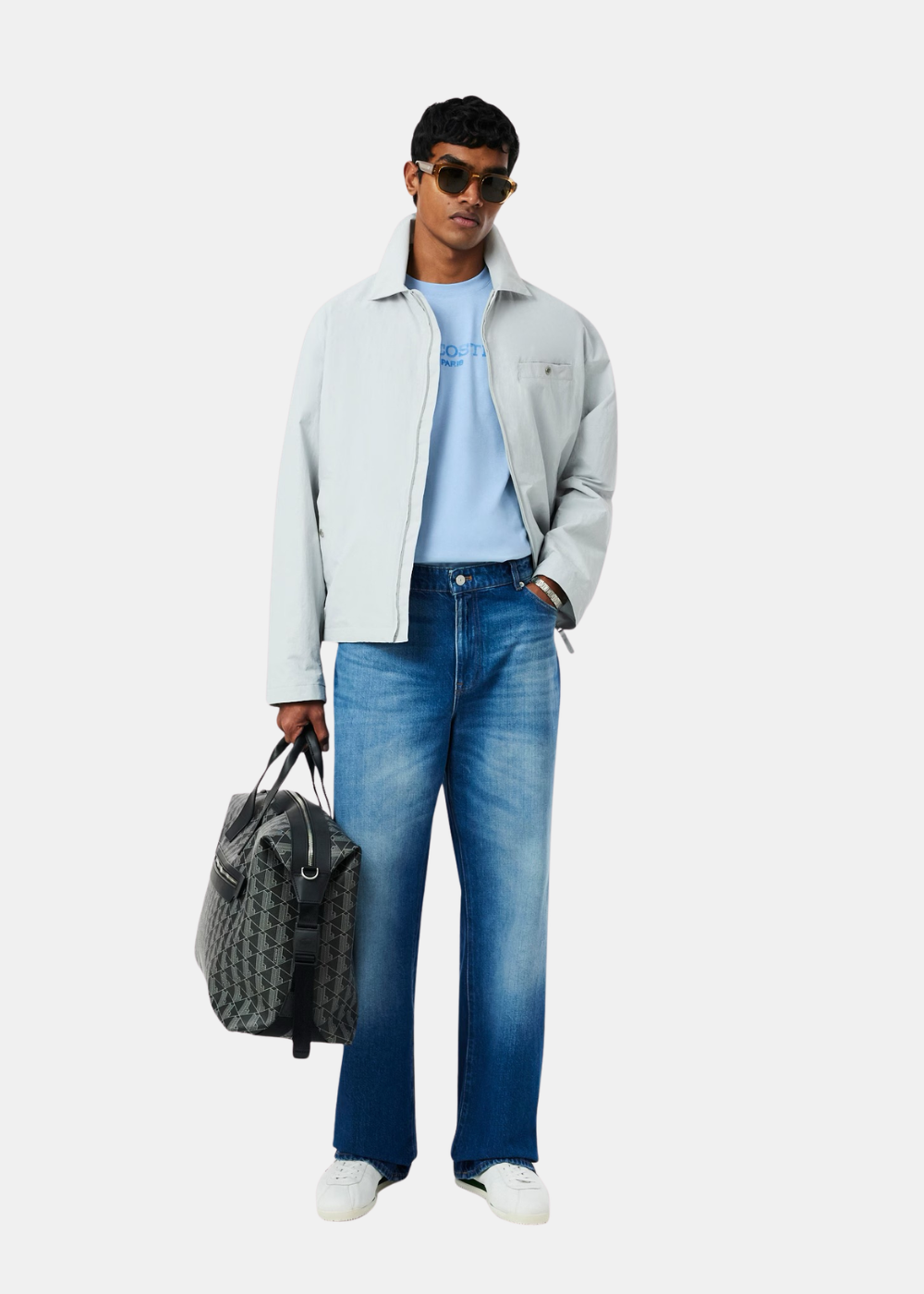 Look complet avec t-shirt bleu ciel Lacoste, veste légère grise, jean bleu, baskets blanches et sac de voyage à motifs