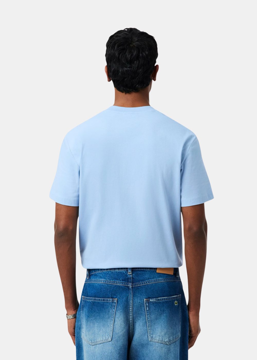 Vue de dos d'un homme portant le t-shirt Lacoste bleu ciel et un jean bleu, mettant en avant la coupe classique du vêtement.