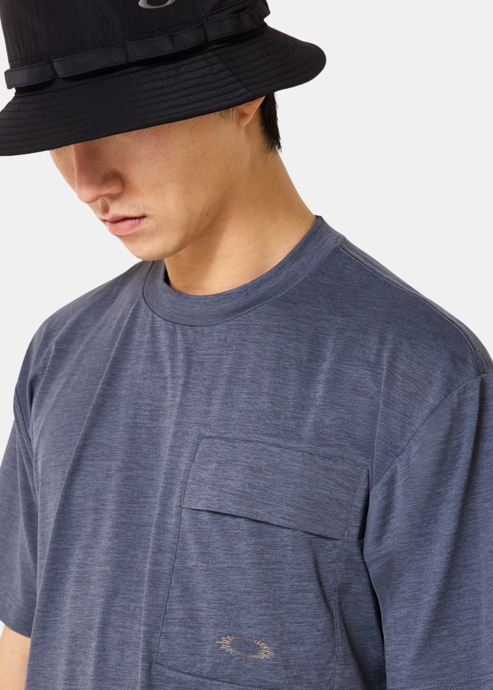 Gros plan sur la poche poitrine à rabat du t-shirt bleu Oakley et la matière chinée, porté avec un bob noir.
