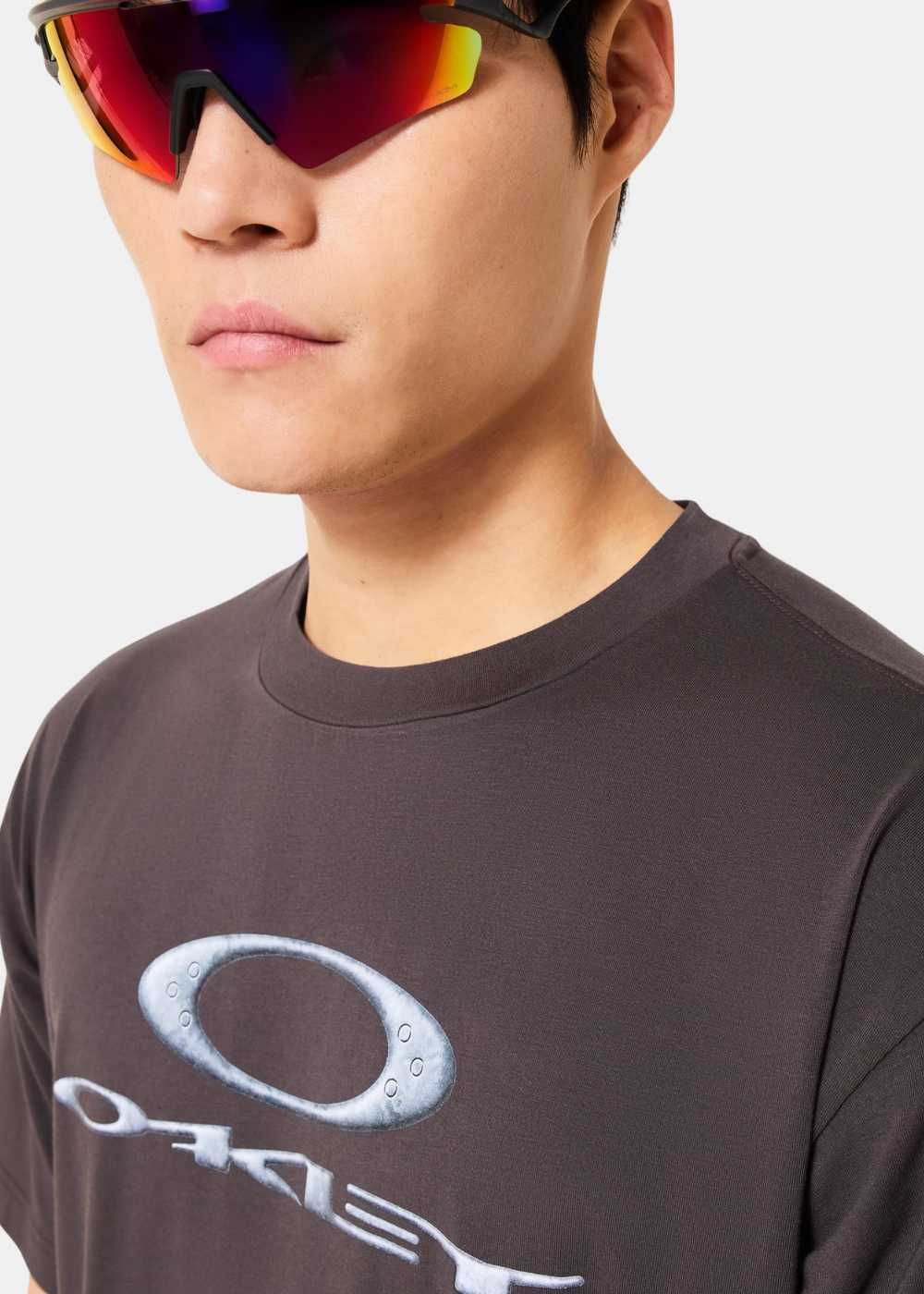 Zoom sur le col et le logo blanc du t-shirt gris Oakley porté par un homme avec des lunettes de sport.