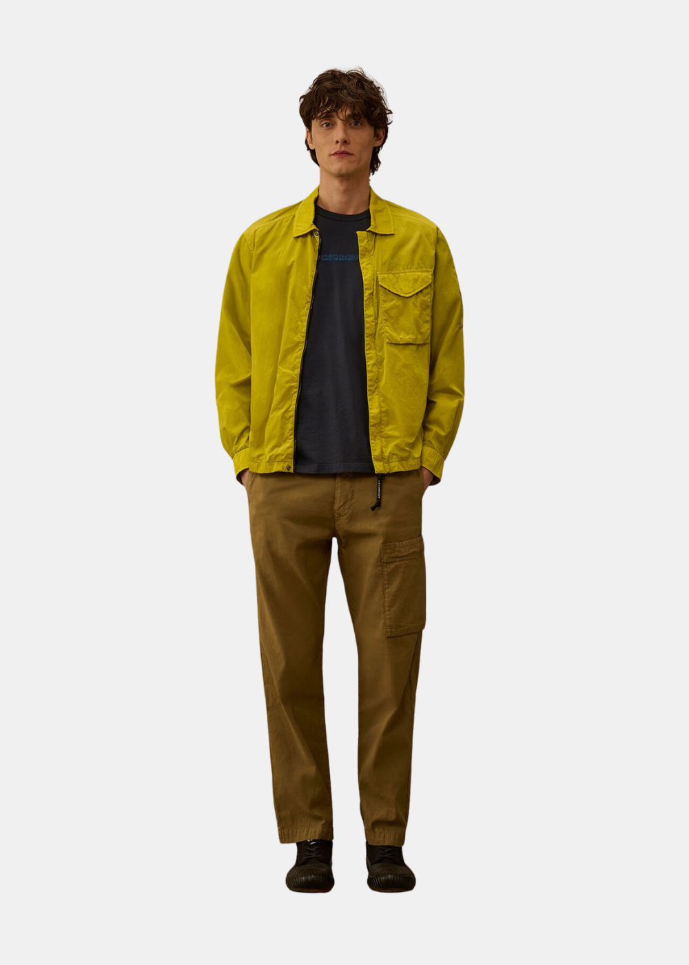 Silhouette en pied du mannequin portant le t-shirt bleu C.P. Company sous une surchemise jaune avec pantalon kaki