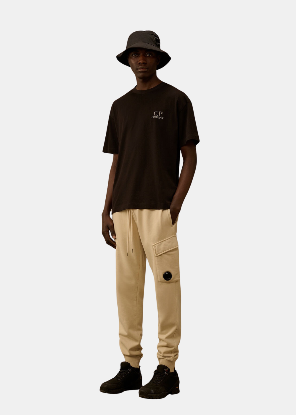 Look complet Homme avec t-shirt noir C.P. Company, pantalon cargo beige et bob noir assorti.