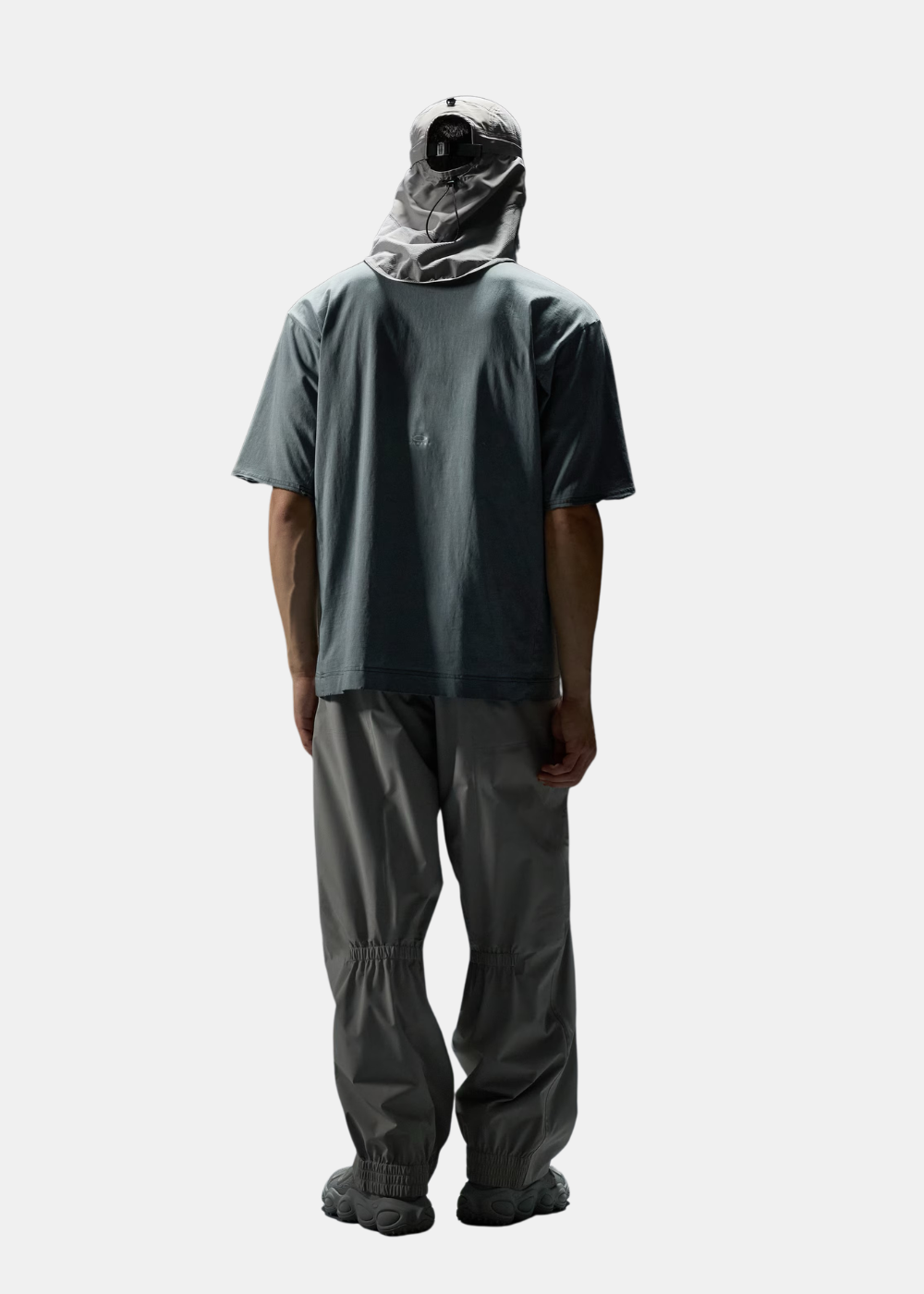 Silhouette complète de dos montrant le t-shirt gris ciment, le pantalon technique et la casquette à rabat.
