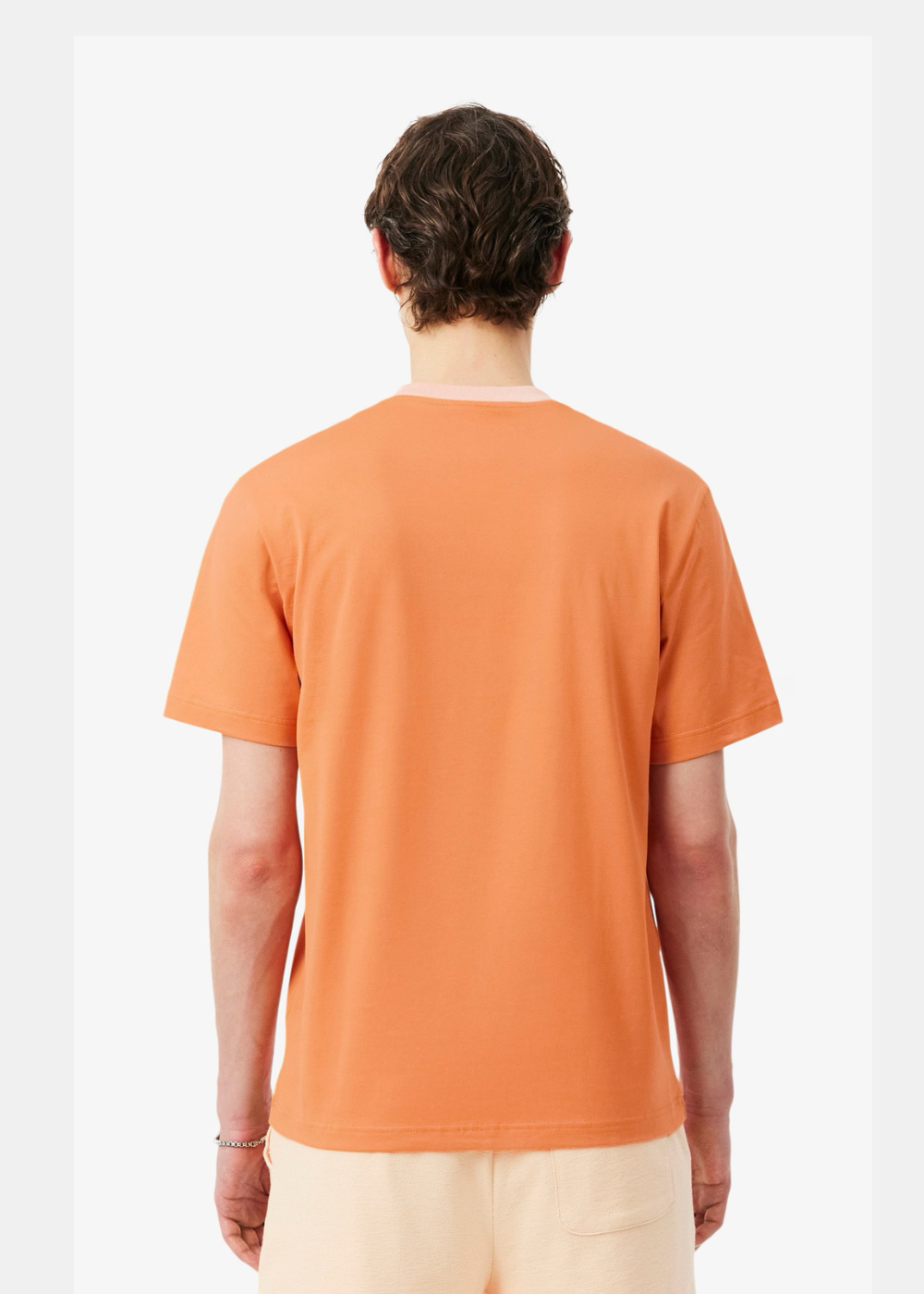 Vue de dos d'un homme portant le t-shirt Lacoste orange vibrant avec le col rose contrasté et un short beige, sur fond blanc.