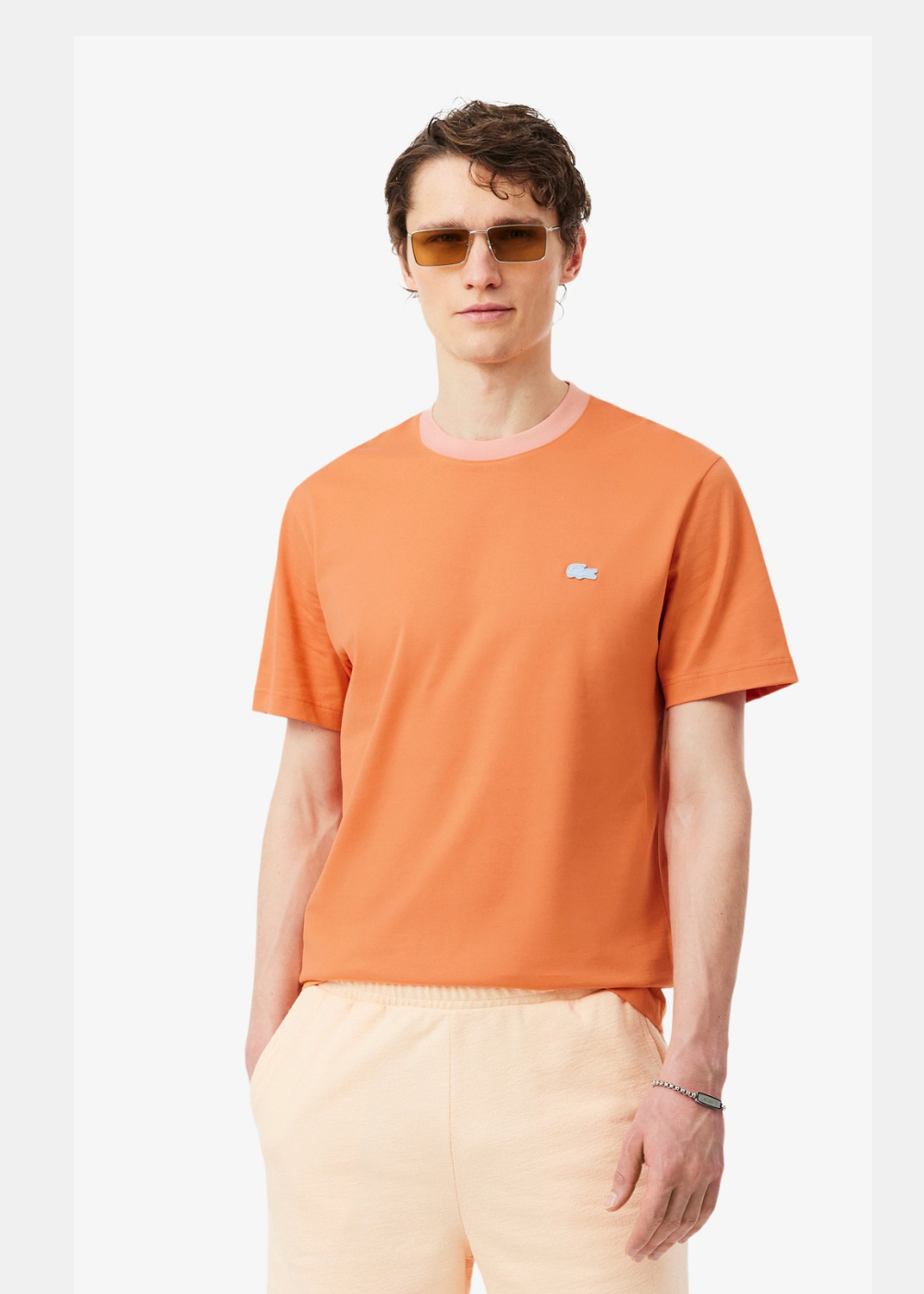 T-shirt Lacoste orange vibrant avec col rose contrasté, porté par un homme avec lunettes de soleil et short beige. Vue de face sur fond blanc.