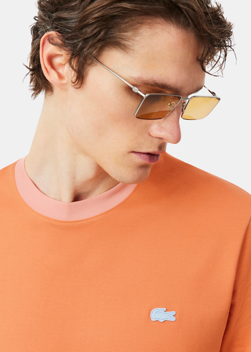 Gros plan sur le buste d'un homme portant le t-shirt Lacoste orange, montrant le col rose contrasté et le logo crocodile bleu brodé.