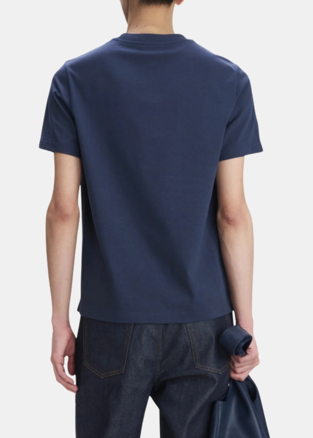 Porté de dos d'un t-shirt marine A.P.C. à coupe droite et col rond, associé à un jean brut, veste tenue à la main.