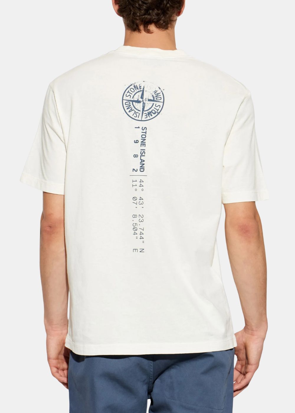 Vue arrière du t-shirt blanc Stone Island Compass porté par un mannequin, affichant la grande boussole bleu marine et l'inscription verticale des coordonnées géographiques au dos.