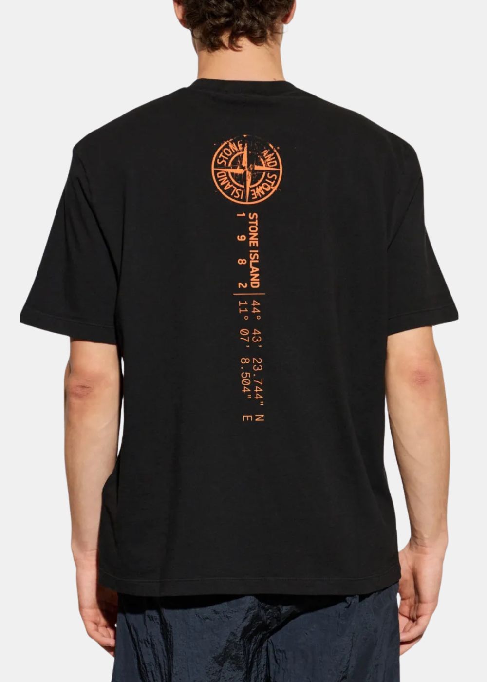 Vue arrière du t-shirt noir Stone Island Compass Coordonnées porté par un mannequin, affichant la grande boussole orange et l'inscription verticale des coordonnées géographiques de la marque au dos.
