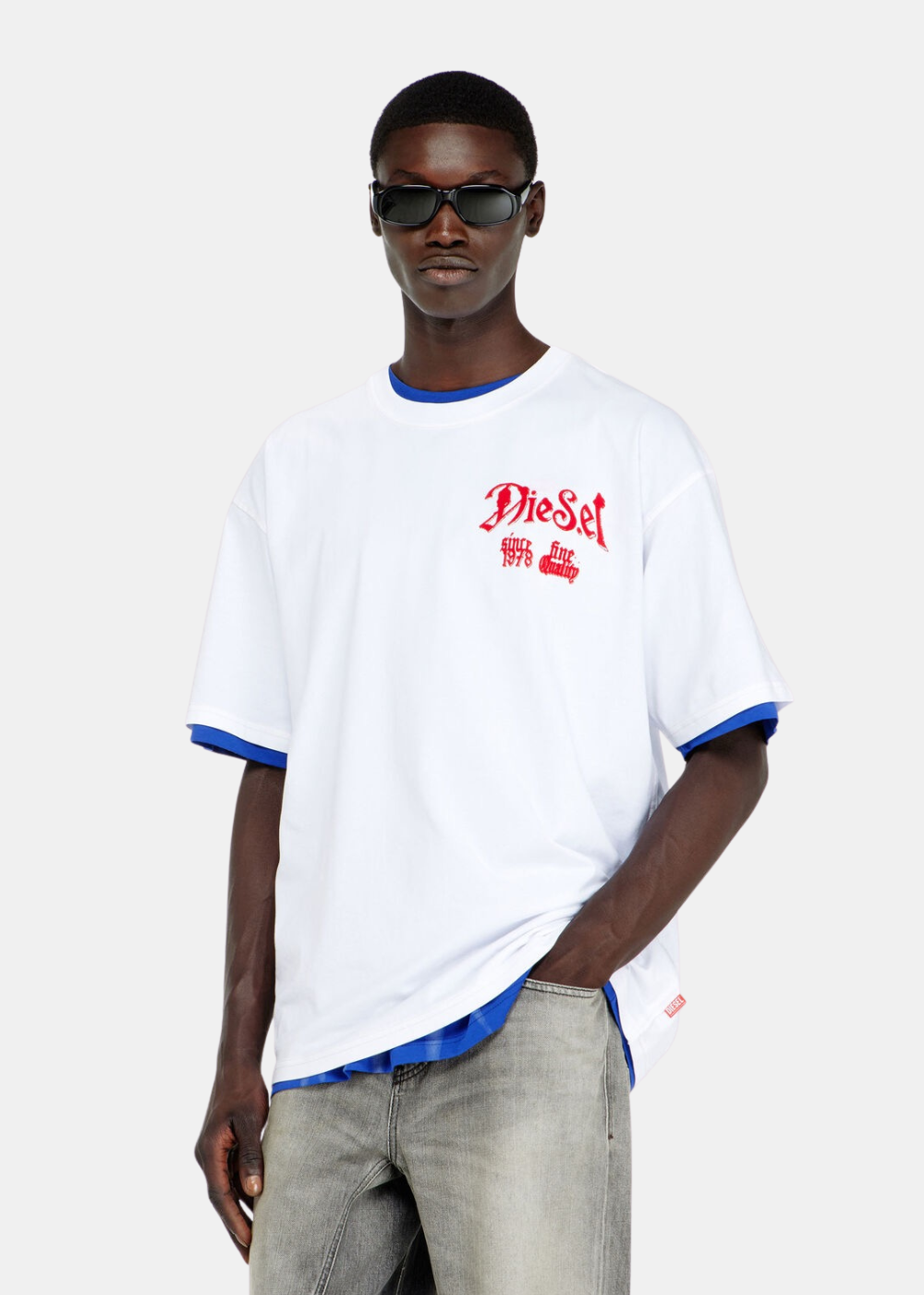 T-shirt Diesel blanc à coupe décontractée porté par un mannequin de face, stylisé en superposition sur un haut bleu et associé à un jean gris clair délavé, mettant en évidence la broderie gothique rouge sur la poitrine