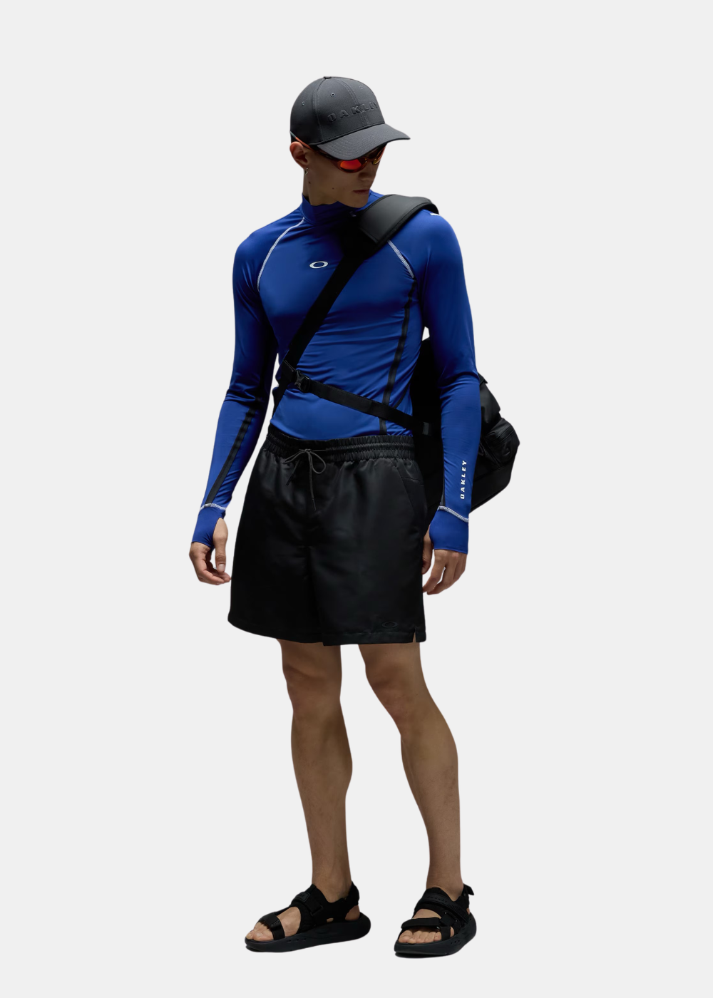 Silhouette entière d'un mannequin équipé du t-shirt manches longues bleu Oakley, d'un short noir, d'une casquette et d'un sac à dos.