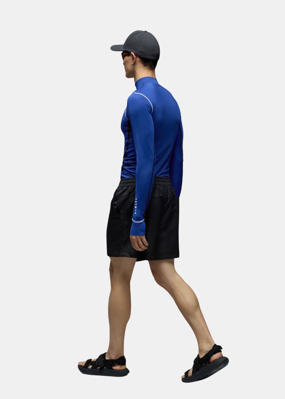 Look complet de profil montrant le t-shirt manches longues bleu Oakley associé à un short noir, une casquette et des sandales.