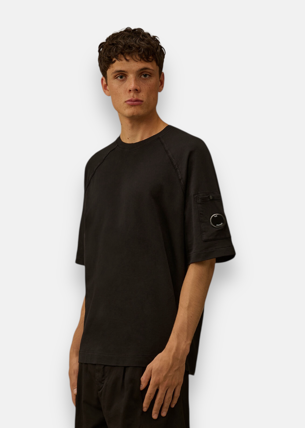 T-shirt noir C.P. Company porté de face par un mannequin mettant en avant la coupe boxy et le logo manche.
