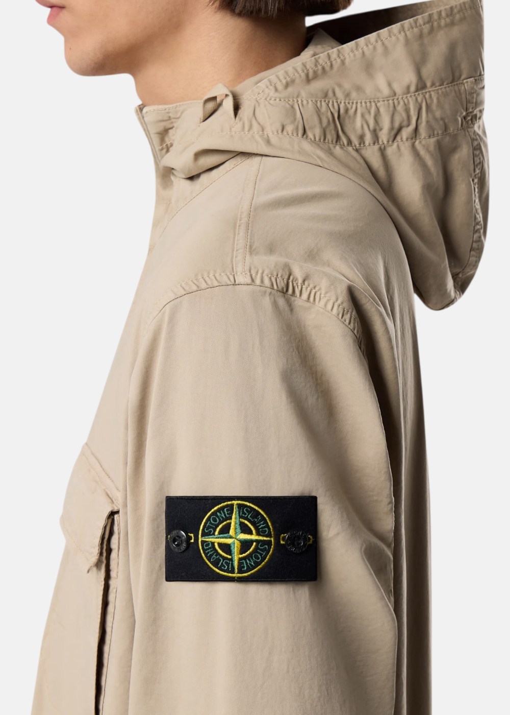 Zoom sur le badge Stone Island sur la manche d'une veste beige avec focus sur la capuche et le profil du mannequin