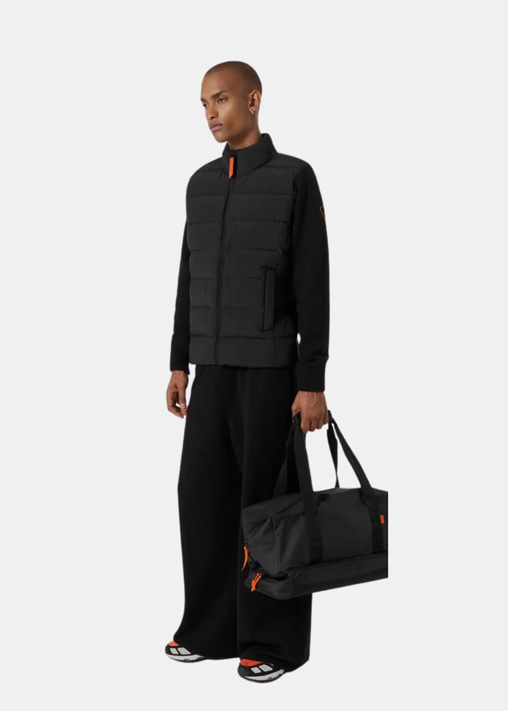 Mannequin homme de face en pied portant la veste hybride matelassée noire Gertrude avec un pantalon ample noir, des baskets et un sac de sport noir