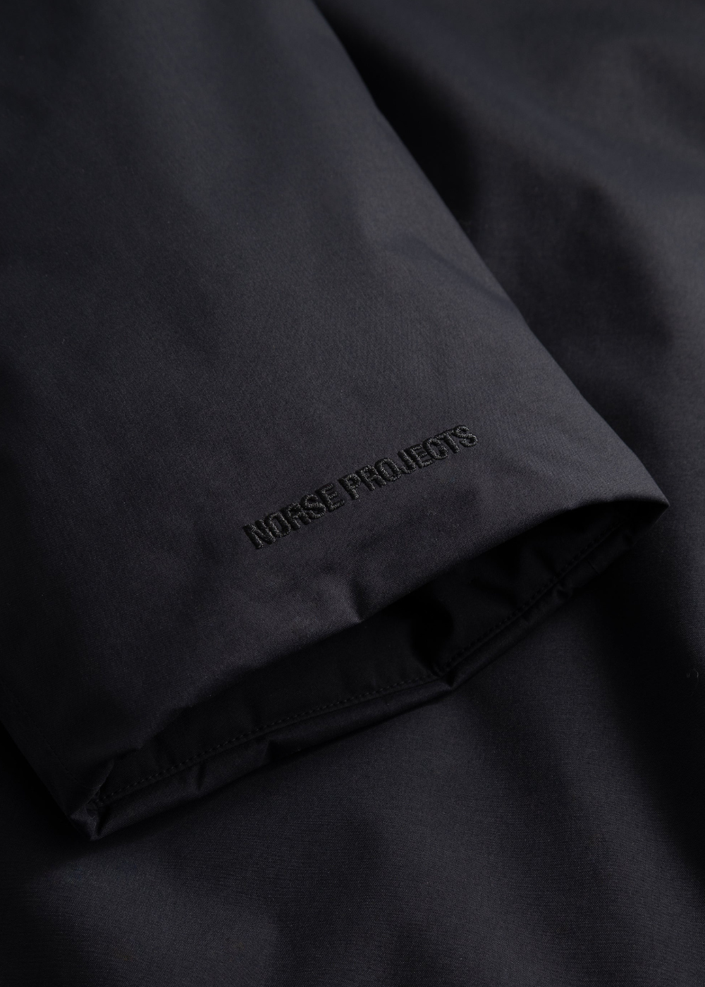 Gros plan sur le bas de manche de la veste Norse Projects noire avec logo brodé ton sur ton
