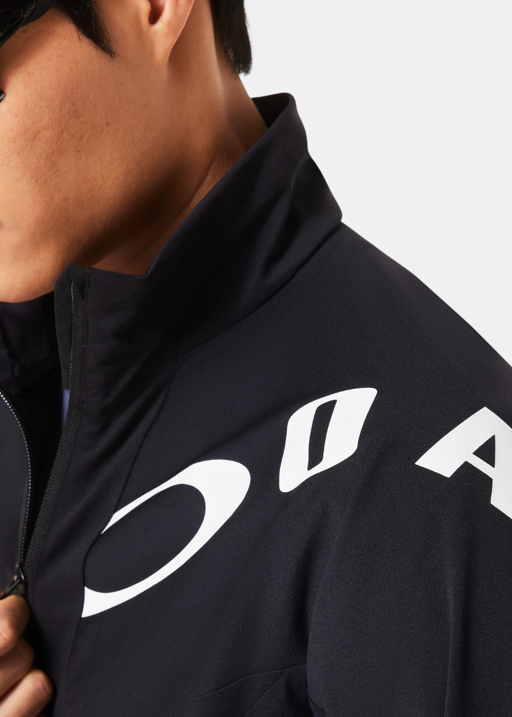 Gros plan sur le col montant et le grand logo blanc imprimé sur l'épaule gauche de la veste noire Oakley.