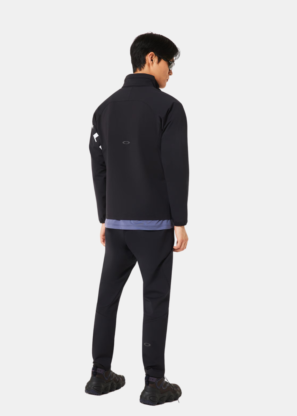Silhouette complète de dos montrant la veste noire Oakley, le pantalon de training assorti et des baskets sombres.