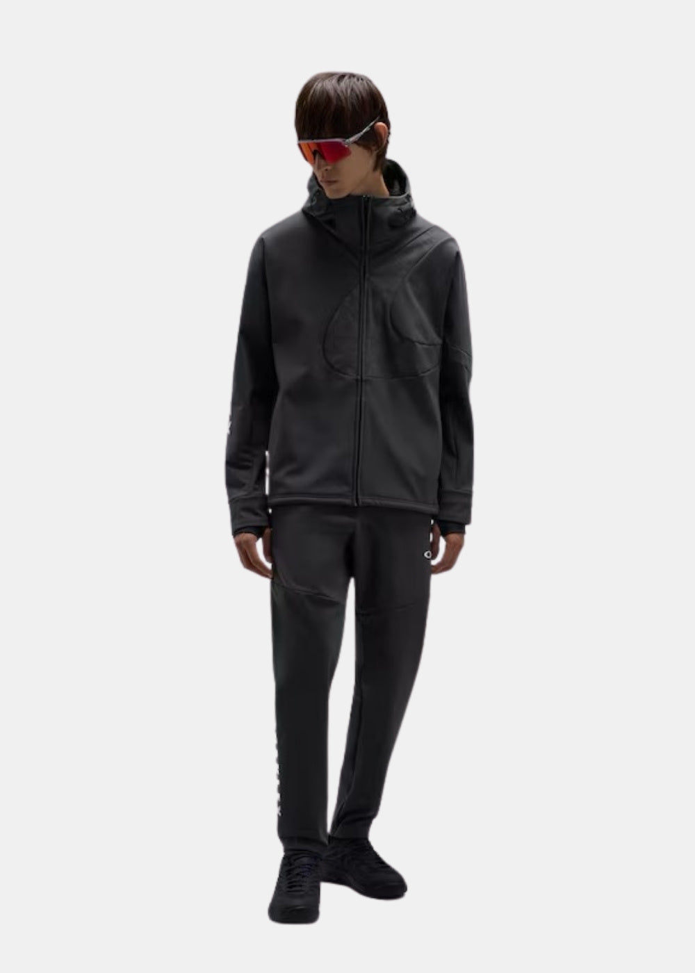 Porté en pied d'une veste technique noire Oakley Enhance à capuche, associée à un pantalon sombre et des chaussures noires.